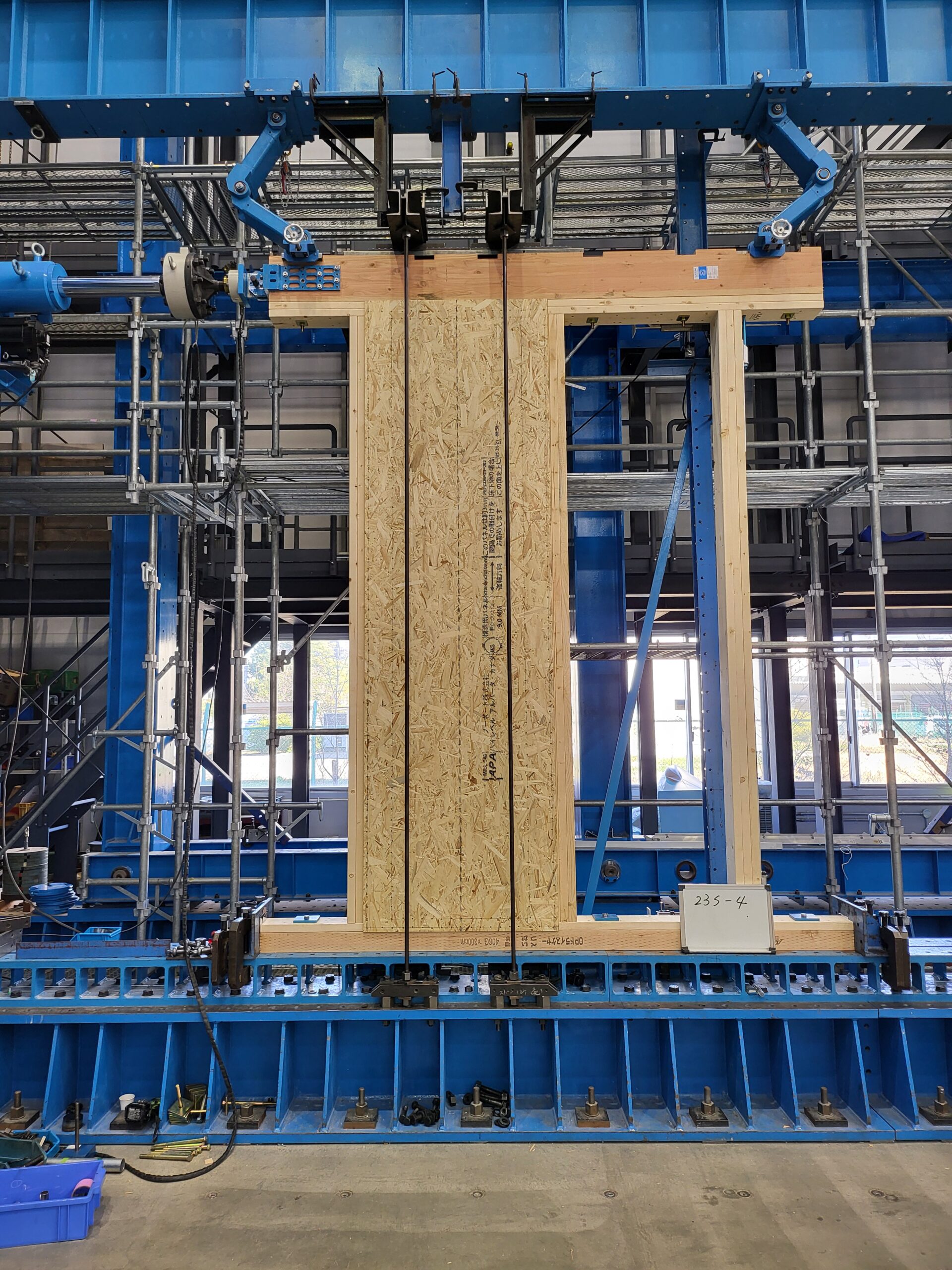 A wooden prefabricated wall component is being held by robotic arms in a research warehouse facility.