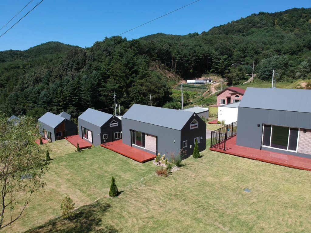 Aerial view of a series of homes on a hill surrounded by lush green trees. The homes have red-painted wood uncovered decks and large windows.