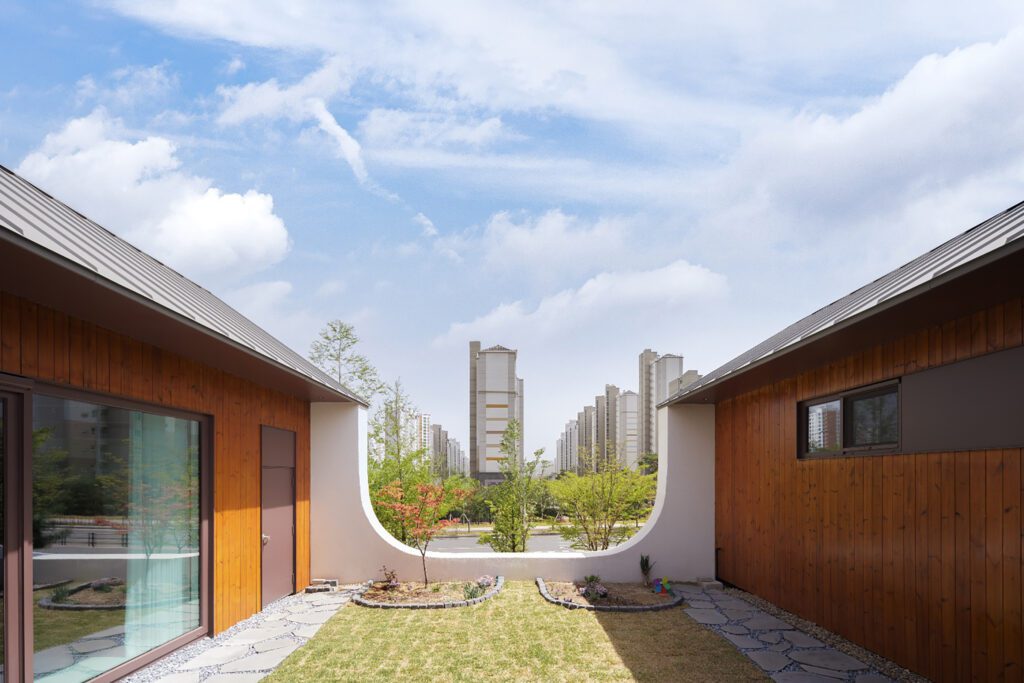 A garden is seen between two homes. A U-shaped wall on the far side of the garden, connects to two homes, while opening the garden up the view to a cityscape of high-rises and a body of water.