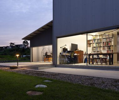 Exterior evening shot of a residence with large glass walls that give view to a room with a desk, dresser, large bookshelves and dining table.