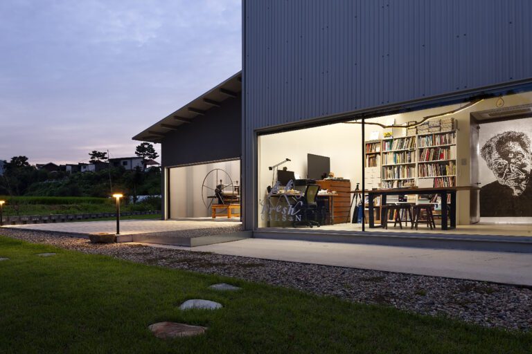 Exterior evening shot of a residence with large glass walls that give view to a room with a desk, dresser, large bookshelves and dining table.